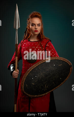 Weibliche Model in Kamera, mit Speer und Schild. Schön, tapfer und ernste Frau in Rot mittelalterlichen Kostüm, Tunika. Frau mit Ingwer Haare stehen, posiert im Studio. Stockfoto