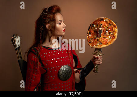 Schöne Frau im mittelalterlichen rote Tunika holding Dolch tragen mit lecker Pizza. Wunderschöne, tapferer Krieger mit Pfeilen hinter zurück, rote Lippen in Studio posieren. Konzept der Junk Food. Stockfoto