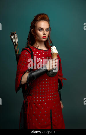 Wunderschöne Modell mit roten Lippen und Ingwer Haar Posing, lecker, weißes Eis. Schöne Frau Krieger tragen in der mittelalterlichen rote Tunika mit Pfeilen hinter zurück, entfernt. Stockfoto