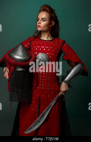 Wunderschöne Frau Krieger Holding mittelalterlicher Dolch und Helm, tragen in Rot mittelalterliche Tunika. Schöne Frau mit roten Lippen und Ingwer Haar in Studio posiert, entfernt. Stockfoto