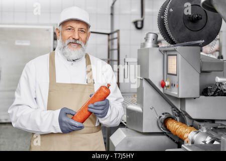 Stattliche Arbeiter in der Nähe von neue, moderne Ausrüstung für Wurstwaren, Würstchen, an der Kamera suchen, posieren. Lebensmittelindustrie. Ältere Mann, der in der weißen Uniform und Schürze. Stockfoto