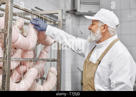 Seitenansicht der älteren Arbeitnehmer tragen in weiße Uniform, braune Schürze und Gummihandschuhe, Arbeiten mit Herstellung von Wurstwaren. Butcher auf modernen Fleisch Manufaktur. Stockfoto