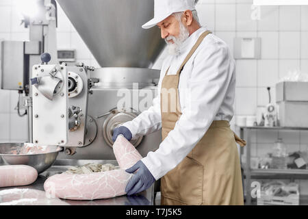 Metzger Mann stand und Arbeiten an Anlagen zur Herstellung von sassages. Ältere Menschen, stattlicher Mann in weißer Uniform und braune Schürze, Gummihandschuhe prüfen und beobachten. Stockfoto