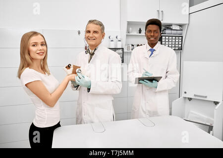 Tierarzt, afrikanische Assistent mit Ordner und Inhaber der Hamster zusammen in Tierklinik posieren. Schöne Frau mit Haustier auf Prüfung. Arzt, Spezialist und Frau lächelnd, mit Blick auf die Kamera. Stockfoto