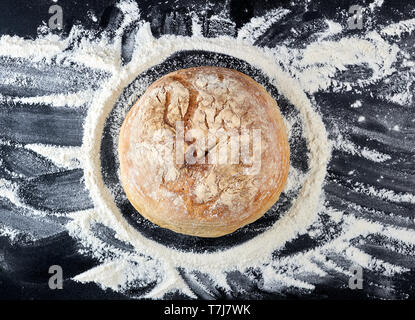 Runde gebackenes Brot und weißes Mehl von Weizen auf einem Schwarzen Tisch verstreut, Ansicht von oben Stockfoto