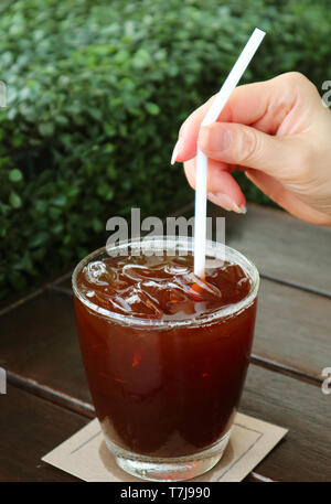 Woman's Hand mit einem Strohhalm in Eiskaffee Glas mit Blurry Green Bush im Hintergrund Stockfoto