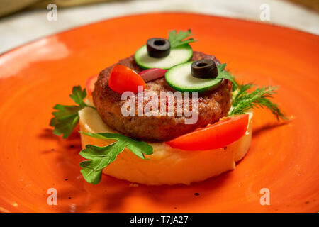 Das Restaurant serviert ein Gericht auf der Speisekarte des Kinder. Kartoffelpüree mit Schnitzel und Gemüse auf eine orange Gesicht in der Form eines Gesichts Stockfoto