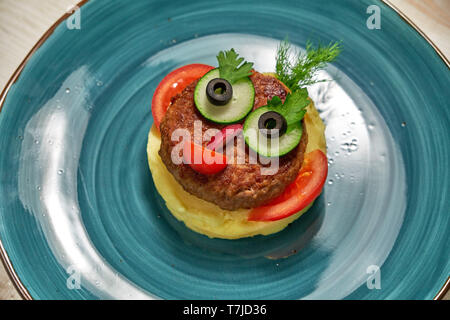 Das Restaurant serviert ein Menü für Kinder auf einem hölzernen Hintergrund. Kartoffelpüree mit Schnitzel und Gemüse auf einem blauen Platte in der Form eines Gesichtes Stockfoto