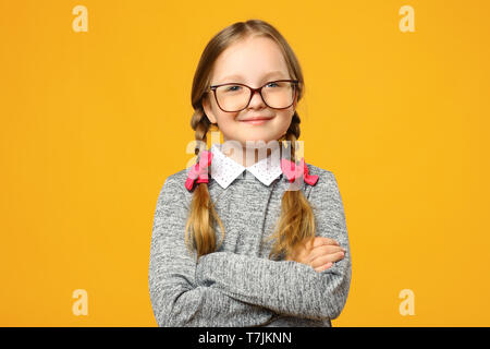 Closeup Portrait von eine lustige kleine Mädchen in Gläsern auf einem gelben Hintergrund. Kind Schulmädchen faltete ihre Arme und sieht in die Kamera. Stockfoto