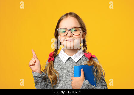 Closeup Portrait kleines Mädchen Schulmädchen auf gelben Hintergrund. Ein Kind von der Grundschule hält ein Lehrbuch und zeigt mit dem Finger nach oben. Das Konzept der Ed Stockfoto