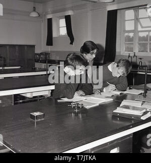 1960, historische, in einer leeren Schule Klassenzimmer, einen weiblichen Mathematik Lehrer beaufsichtigt die Studie oder buchen Sie Arbeit von zwei männlichen Schüler, die mehr studieren oder Überarbeitung nach Klasse, South London, England, UK. Stockfoto