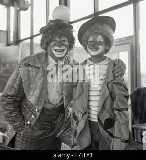 1960, historische, alle lächeln... Spaß in der Schule, ein Zirkus Clown vistin eine Grundschule für ein Foto neben einem lächelnden jungen Schüler in Fancy Dress und mit seinem Gesicht wirft als Clown, England, UK gemalt. Stockfoto