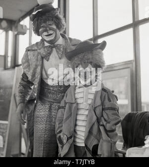 1960, historische, einem Zirkus Clown besuchen eine Grundschule mit einem jungen Schüler tragen Fancy Dress und mit seinem Gesicht als Clown, England, UK gemalt. Stockfoto