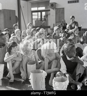 1960, historische, junge Schülerinnen und Schüler in der Aula zusammen sitzen auf dem Boden lachen, durch den Gastredner unterhält, ein Zirkus Clown, England, UK. Stockfoto