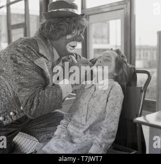 1960, historische, einem Zirkus Clown, Gesicht Farbe auf einer aufgeregt, aber etwas ängstliche junge Schulmädchen, England, UK. Stockfoto