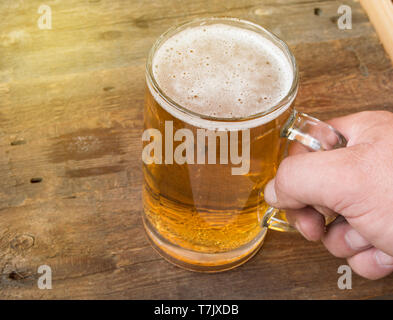 Sommer, Party, des Menschen Hand mit einer Tasse helles Bier auf dunklen alten hölzernen Brettern. Stockfoto