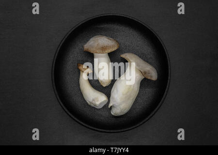 Abstrakte Komposition von drei Eringi Pilze in schwarz Gusseisen Schüssel auf grauem Stein Hintergrund Stockfoto