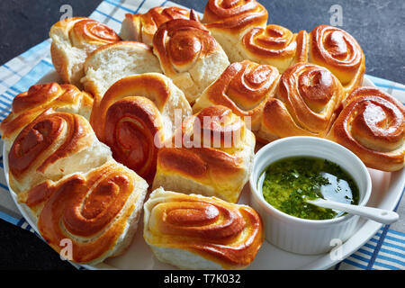 Close-up Ziehen neben Brötchen, hausgemachter Hefe Abendessen Brötchen auf einer weißen Platte mit Knoblauch Petersilie in einer Schüssel auf einer konkreten Tabelle, horizontale Ansicht Stockfoto