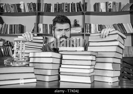 Lehrer oder Schüler mit Bart sitzt am Tisch mit Büchern, defokussiert. Mann auf ernstes Gesicht zwischen den Stapeln von Büchern, während ihres Studiums in Bibliothek, Bücherregale für den Hintergrund. Bibliophile Konzept. Stockfoto