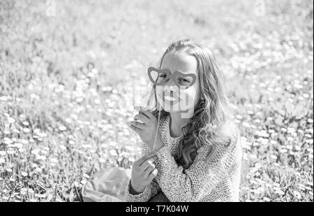 Liebe auf den ersten Blick. Mädchen auf Happy Face verbringen Freizeitaktivitäten im Freien. Mädchen sitzt auf Gras an grassplot, grünen Hintergrund. Kind mit Karton Herzform Brillen und lächelnden Mund posieren. Stockfoto