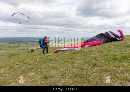 Devils Dyke, Sussex, UK; 6. Mai 2019; Männliche Gleitschirm steht neben seinem Stürzte ein Baldachin. Drei Gleitschirme fliegen im Hintergrund Stockfoto