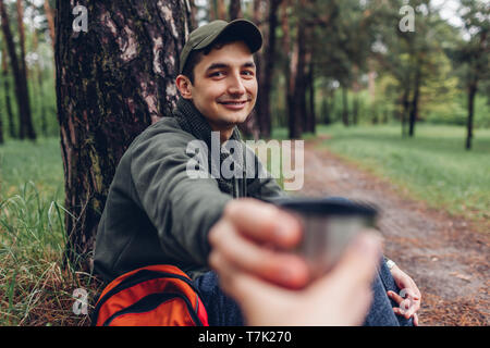 Junge glücklicher Mann touristische geben heiße Tasse Tee an seinen Freund im Frühjahr Wald. Camping, Reisen und Sport Konzept Stockfoto