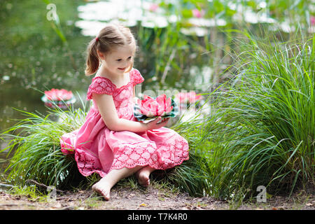 Kind am Seeufer sitzen beobachten Seerose Blüten. Kleines Mädchen mit rosa Lilie Blume. Zicklein an Lilien auf sonniger Frühlingstag. Kinder outdoor Stockfoto