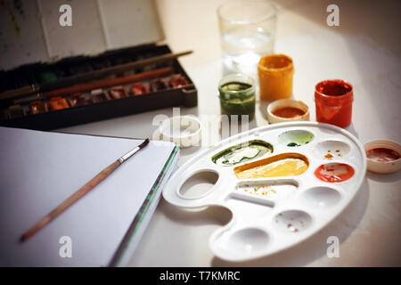 Auf dem Tisch gibt es Aquarellfarben in einem Jahrgang, ein großes Notebook zum Zeichnen auf eine Feder, ein Glas Wasser für Waschbürsten, offene gouach Stockfoto