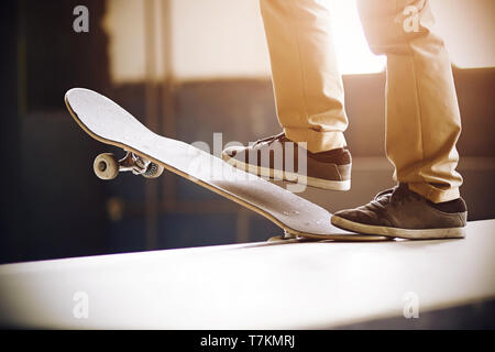 Ein Kerl in beige Hosen und schwarze Turnschuhe steht auf einer Rampe auf einem Skateboard und einem Rückgang der Trick, durch die freudige Sonnenlicht beleuchtet Stockfoto