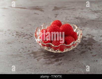 Seitenansicht des frisch gepflückte Himbeeren in eine kleine Glasschüssel auf grauem Hintergrund mit natürlichem Licht beleuchtet. Stockfoto