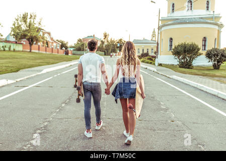 Ein junges Paar Wanderungen im Sommer in der Stadt, in den Händen von skateboards, longboard. Ansicht von hinten. Spaziergang am Wochenende, Lebensstil der jungen Menschen. Stockfoto