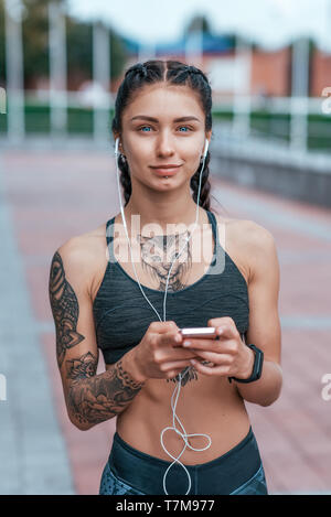 Schöne Frau lächelnd, glücklich im Sommer in der Stadt, gebräunte Haut tattoo. Sportswear top. Telefon Ohrhörer. Konzept der Kraft, Weiblichkeit, Mut Stockfoto