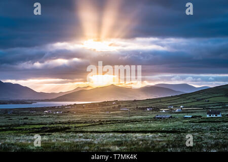 Sonnenaufgang über der Halbinsel Dingle, betrachtet aus Ring of Kerry. Reetdachhaus im Vordergrund, Strahlen der Sonne durch die Wolken. Stockfoto