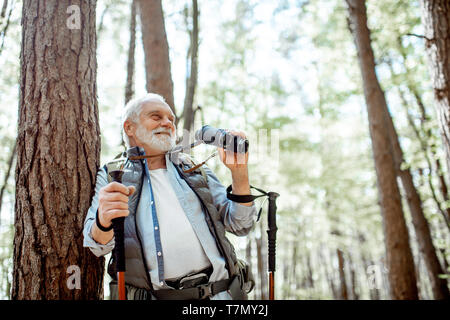Portrait eines älteren Mannes mit Fernglas und Rucksack ruht in der Nähe des Baumes Unterwegs im Wald Stockfoto