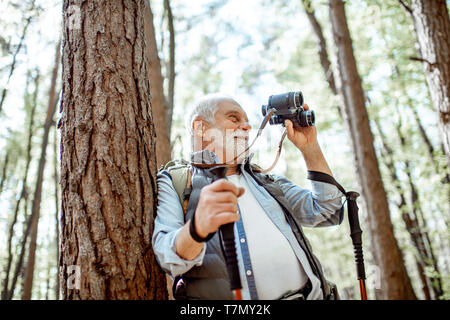 Portrait eines älteren Mannes mit Fernglas und Rucksack ruht in der Nähe des Baumes Unterwegs im Wald Stockfoto