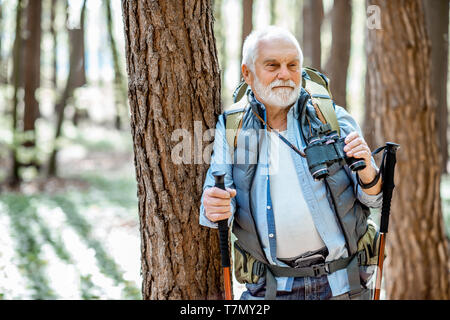 Portrait eines älteren Mannes mit Fernglas und Rucksack ruht in der Nähe des Baumes Unterwegs im Wald Stockfoto