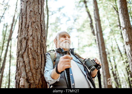 Portrait eines älteren Mannes mit Fernglas und Rucksack ruht in der Nähe des Baumes Unterwegs im Wald Stockfoto