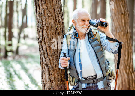 Portrait eines älteren Mannes mit Fernglas und Rucksack ruht in der Nähe des Baumes Unterwegs im Wald Stockfoto