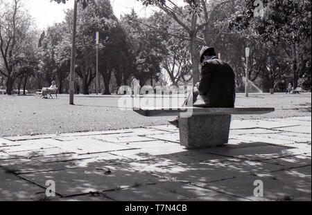 1977 Sydney, Australien: ein Mann im Anzug und Trilby Hut sitzt auf einem modernen Design preform konkrete Sitz in dappled Schatten im Hyde Park. Im Hintergrund andere Leute durch den Park zu Fuß oder auf eher traditionelle Holz und Stahl in der warmen Sonne zu sitzen. Stockfoto