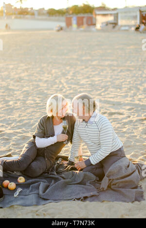 Ältere Frau und älterer Mann sitzen auf Plaid auf Sand Strand und trinken Champagner. Konzept der Rentner auf Picknick. Stockfoto