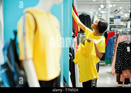 Afican amerikanische Frauen in Trainingsanzügen einkaufen bei Sportswear Mall gegen Regale. Sie wählen Gelb t-shirt. Sport store Thema. Stockfoto