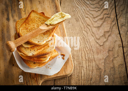 Holz- butter Spreaders auf einem Stapel von knusprigem golden weiß Brot Toast auf einem Holzbrett über einen rustikalen Holztisch mit Holzmaserung Muster und kopieren Raum Stockfoto