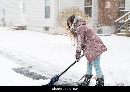 Junge Frau im Winter Mantel aus Schnee schaufeln Fahrstraße Straße in schweren Schneesturm mit Schaufel von Wohnhäusern im Hinterhof Stockfoto