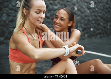 Lächelnden jungen Frau in Sportkleidung Kontrolle ihr Sport ansehen, während sie auf dem Bürgersteig sitzen mit ihrem Lachen Freund nach einem Durchlauf zusammen Stockfoto