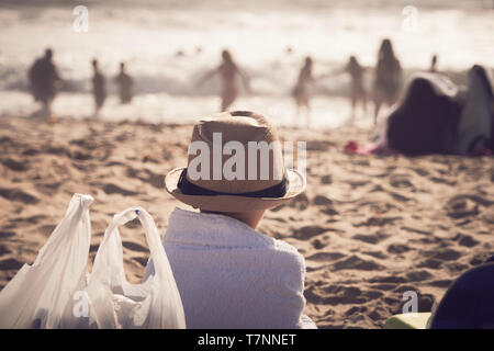 Junger Mann, Hut und in Handtuch gewickelt sitzt auf Sandstrand und Uhren Leute spielen im Wasser in San Diego, Kalifornien. Urlaub Sommer, Reisen, entspannen. Stockfoto