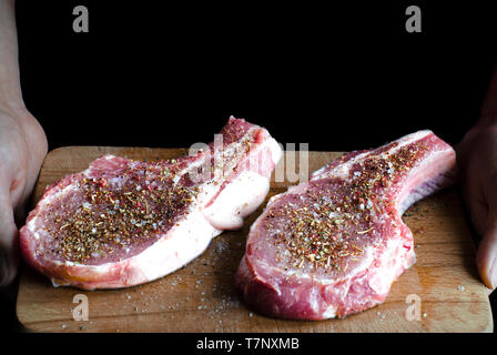 Hände halten einen Vorstand, an dem zwei rohes Schweinefleisch Steaks mit Gewürzen und bereit zu Kochen bestreut sind. Raw marmoriertes Fleisch Steak, Pfeffer, Kräuter, alte hölzerne Stockfoto