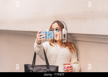 Porträt der Schönen attraktiven jungen trendigen Mädchen in Pullover und Jeans mit Kopfhörern, Sonnenbrille und Smart Phone nimmt selfie während lehnte sich an der Wand Stockfoto