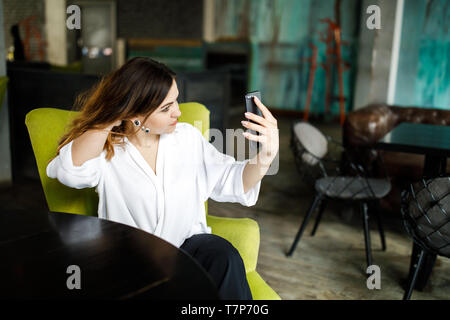 Eine junge, sympathische Frau, nicht eine dünne - vorangegangen Body Building, sitzt in einem gemütlichen Cafe, hält ein Smartphone in den Händen und hebt sich. Stockfoto