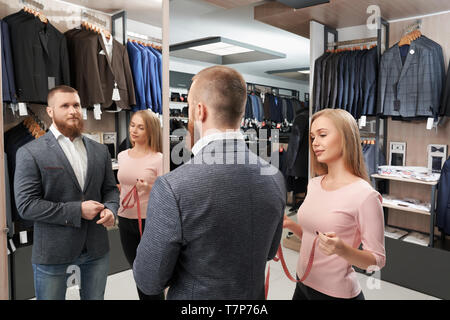 Ansicht von der Seite der bärtige Mann, der versucht, die neuen weißen Hemd und grau casual Jacke im Shop. Lächelnder Mann, der vor Spiegel, während die weiblichen maßgeschneiderte Messung von Länge der Hülse. Konzept der Armatur. Stockfoto