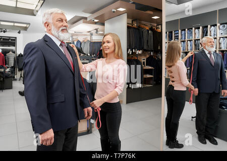 Seitenansicht des eleganten ältere Mann, der versucht, auf neue formale Klage in der Boutique. Stilvolle männlichen Klienten zu spiegeln, während die weiblichen assistant Messung Länge auf dem Ärmel. Konzept der Kleidung und die Montage. Stockfoto